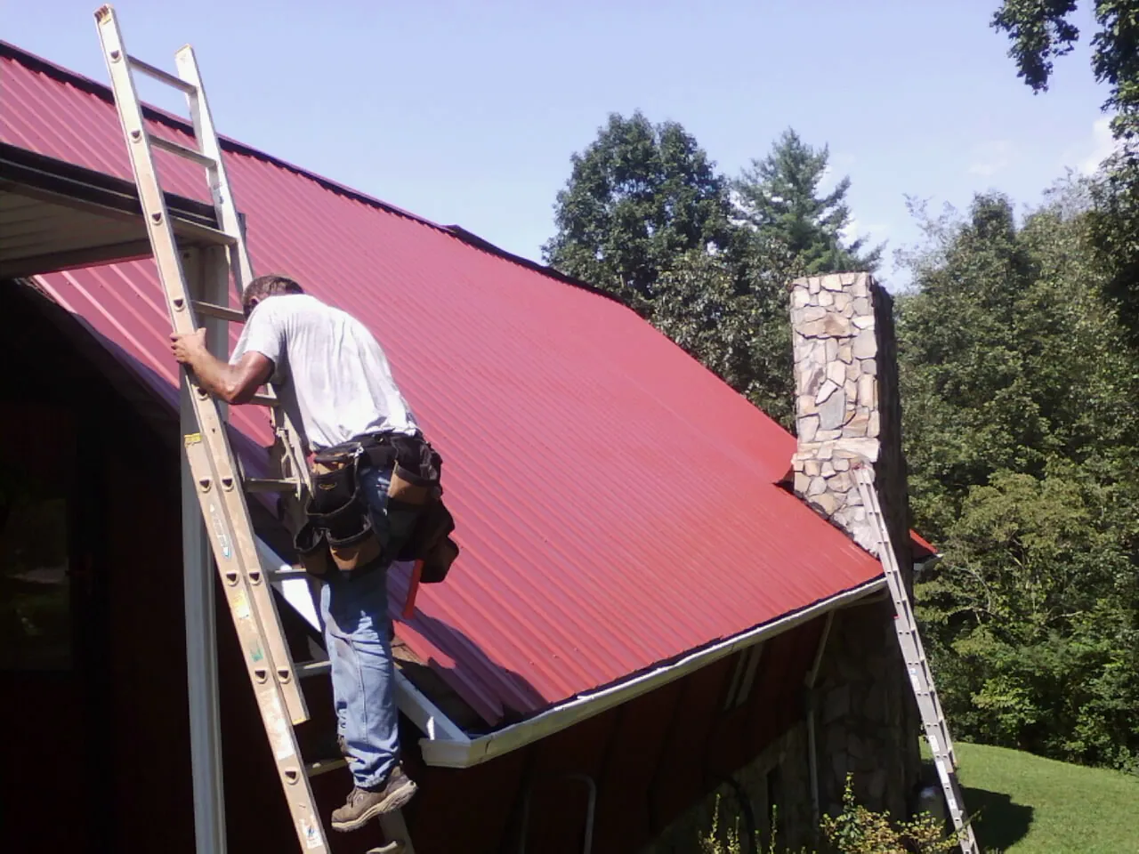 Roofer on a ladder beside a red metal roof and stone chimney — Soffit & Fascia Repair by Roofing Brothers in Yucaipa